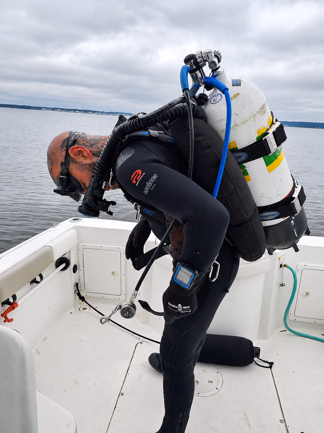 Diver in black wetsuit and rebreather gear bending over on a boat near calm water under cloudy sky.