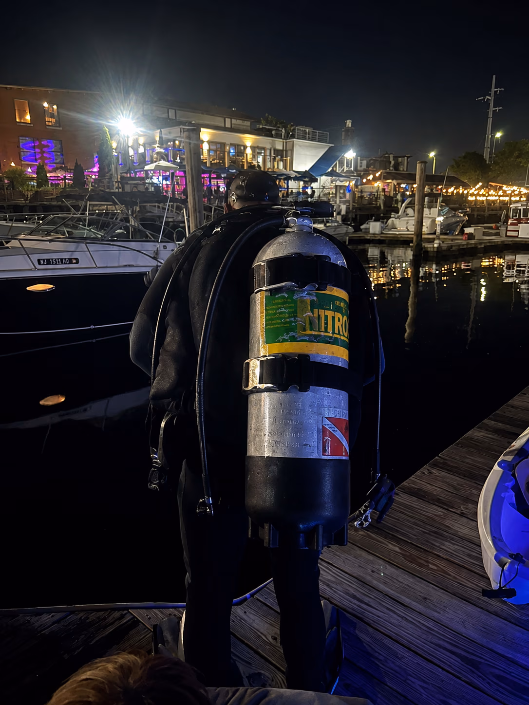 Scuba diver in black wetsuit with nitrox tank standing on a dock by boats at night.