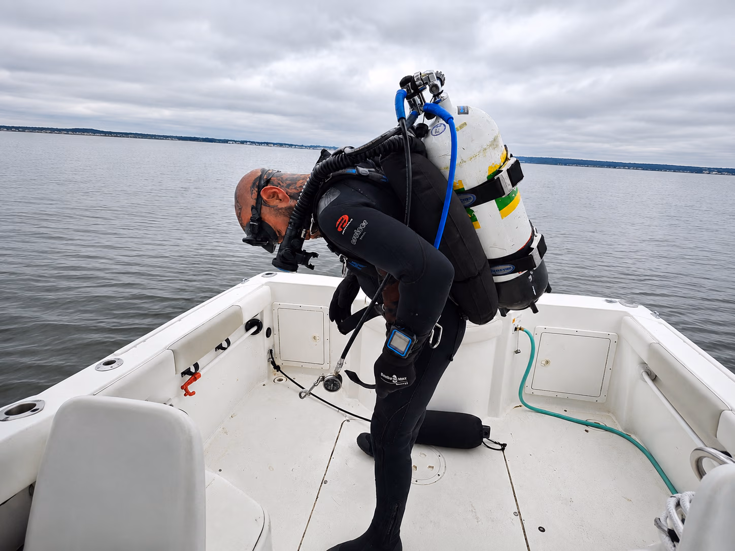 Scuba diver in wetsuit with tanks bends over on a boat with cloudy sky and water background.