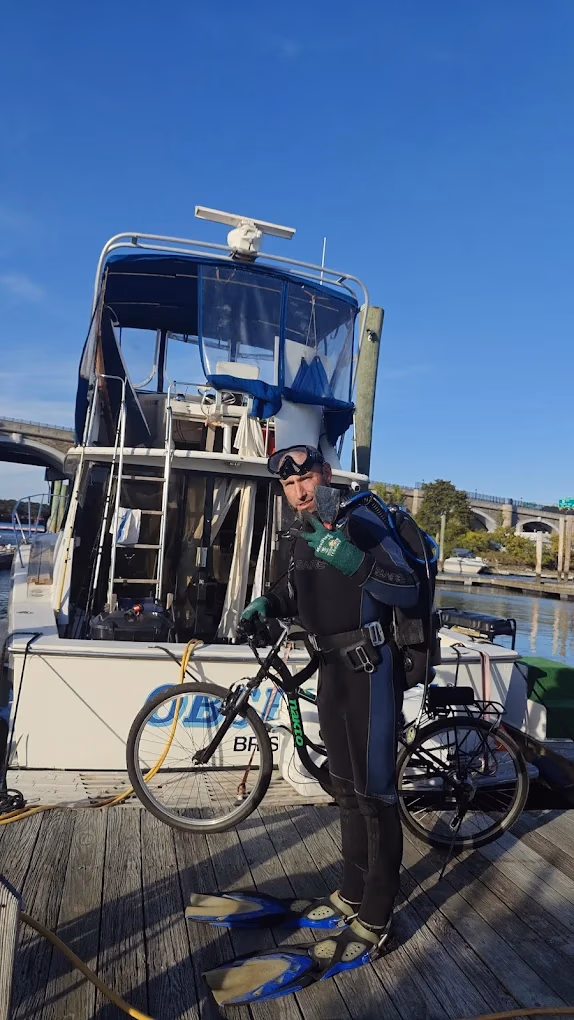 Man in scuba gear holding a bicycle on a dock next to a moored boat under blue sky.