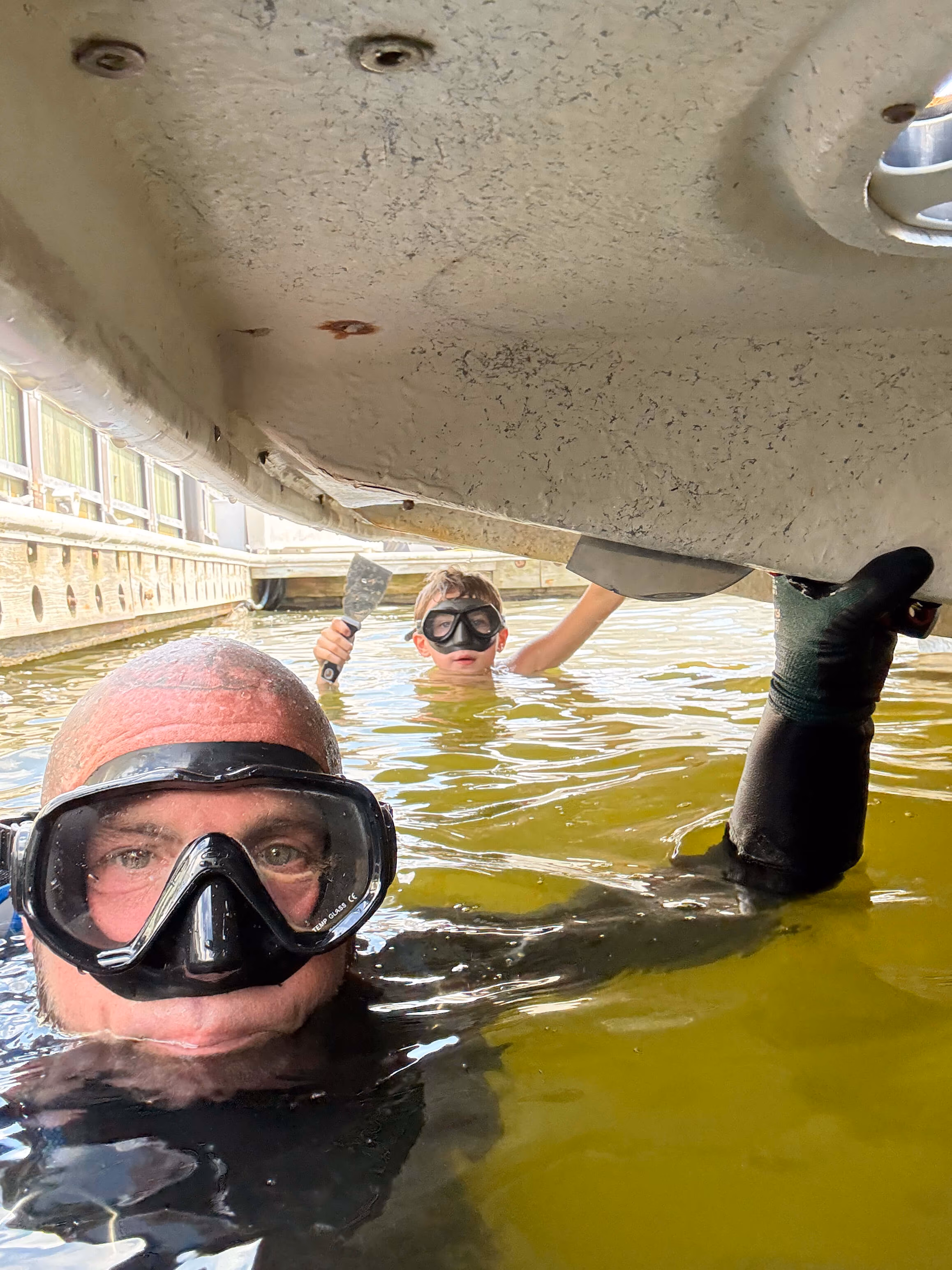Two divers wearing masks, one holding a putty knife, working under a boat in greenish water near a dock.