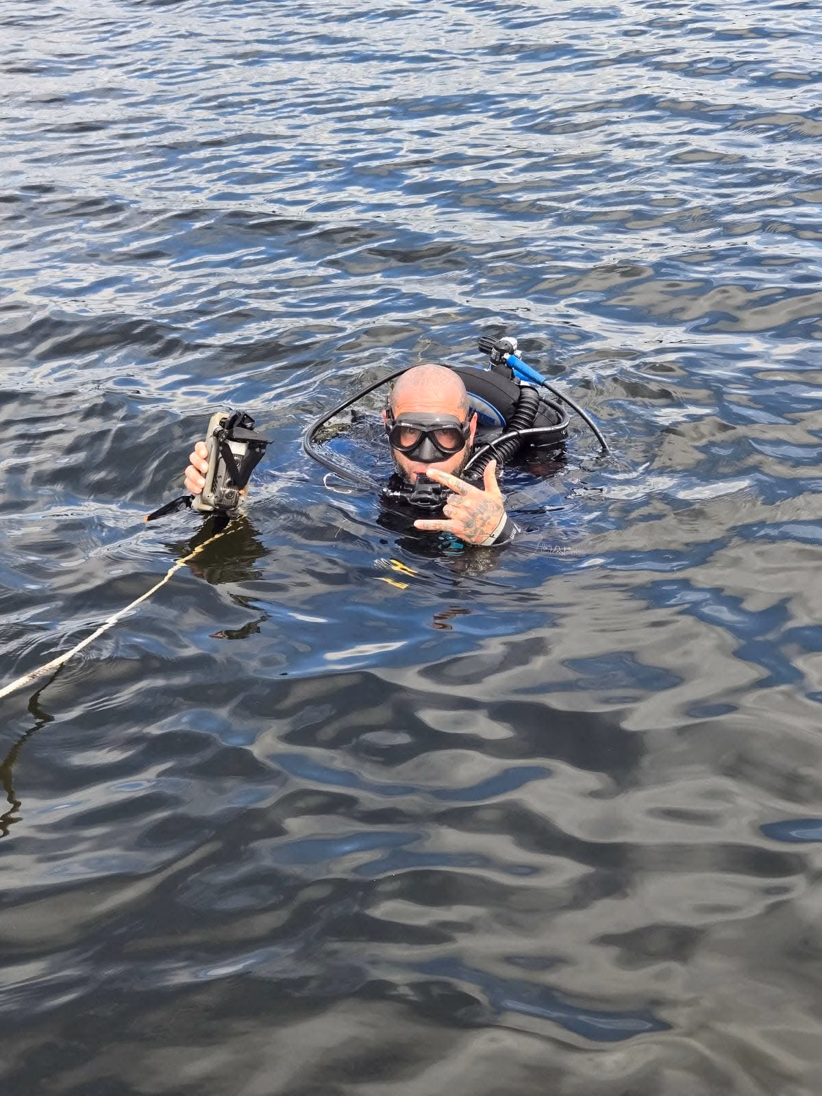 Diver with scuba gear holding a camera and making a hand gesture in rippled water.