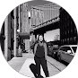 Man in dark clothing sits on street curb beside parked cars under an urban bridge.