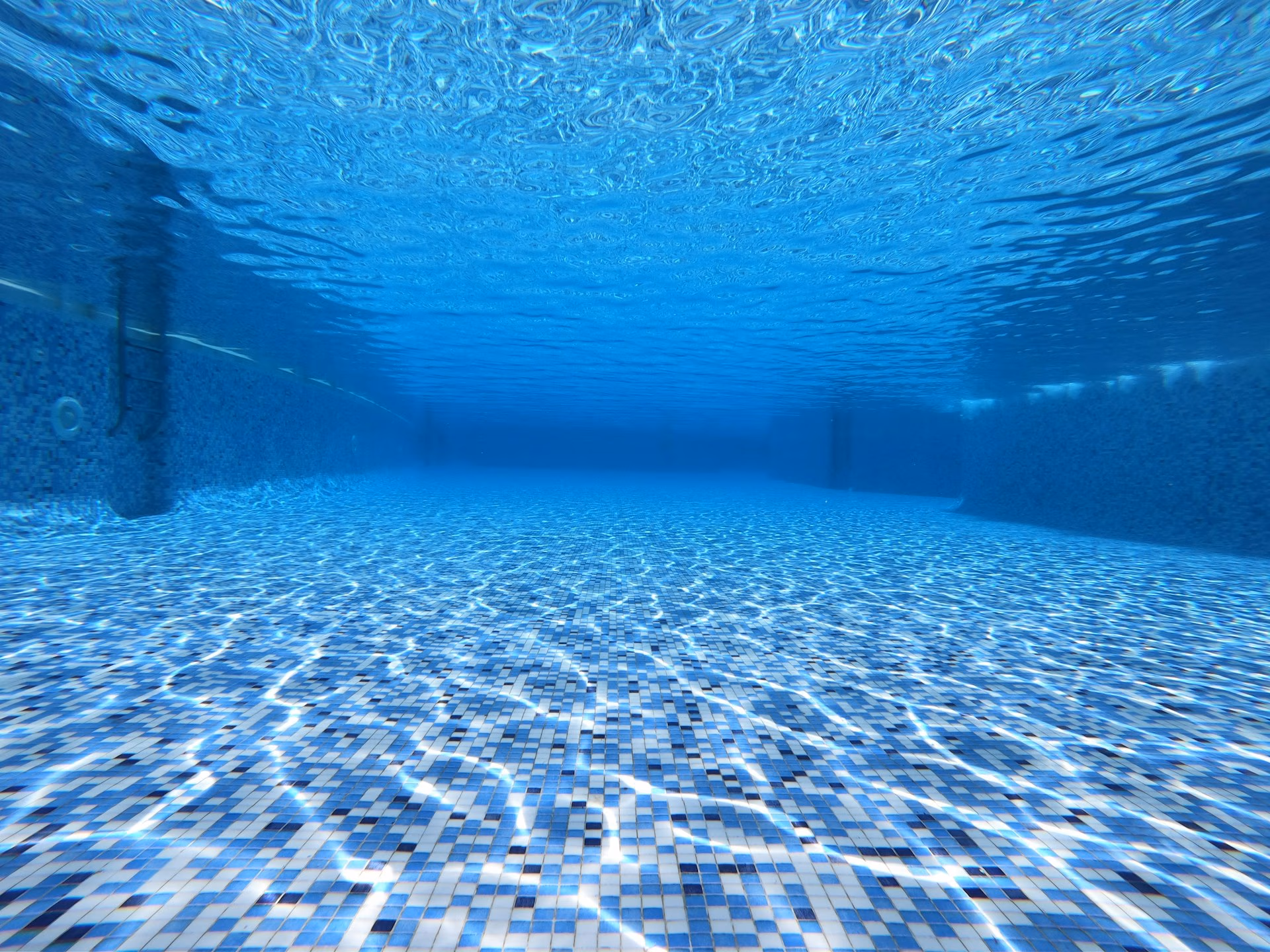 Underwater view of an empty swimming pool with blue mosaic tile flooring and walls.