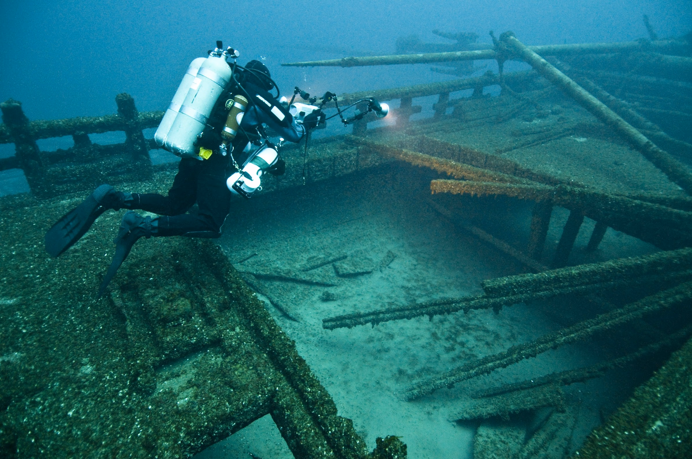 Scuba diver with camera and lights exploring a barnacle-covered shipwreck underwater.